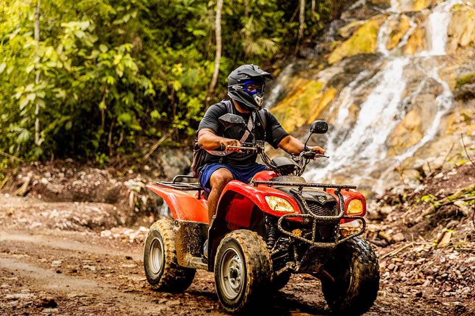 A rider on a red quad pausing on a muddy track beside a waterfall running down the rock face.