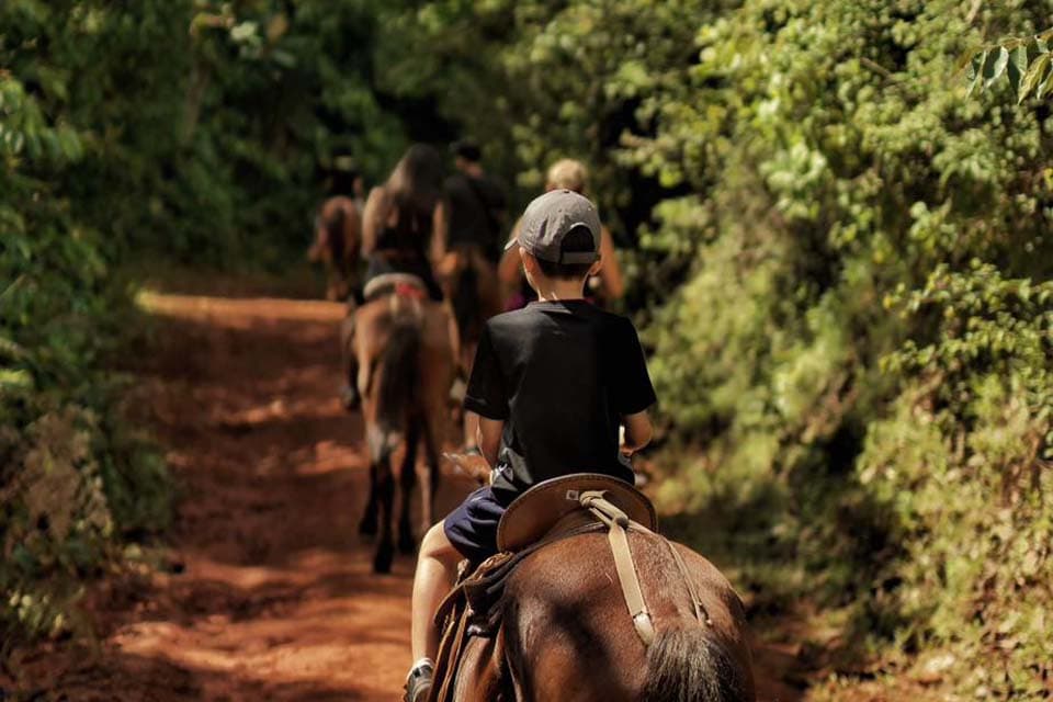 A child in a baseball cap riding at the back of the line up a red-dirt track.