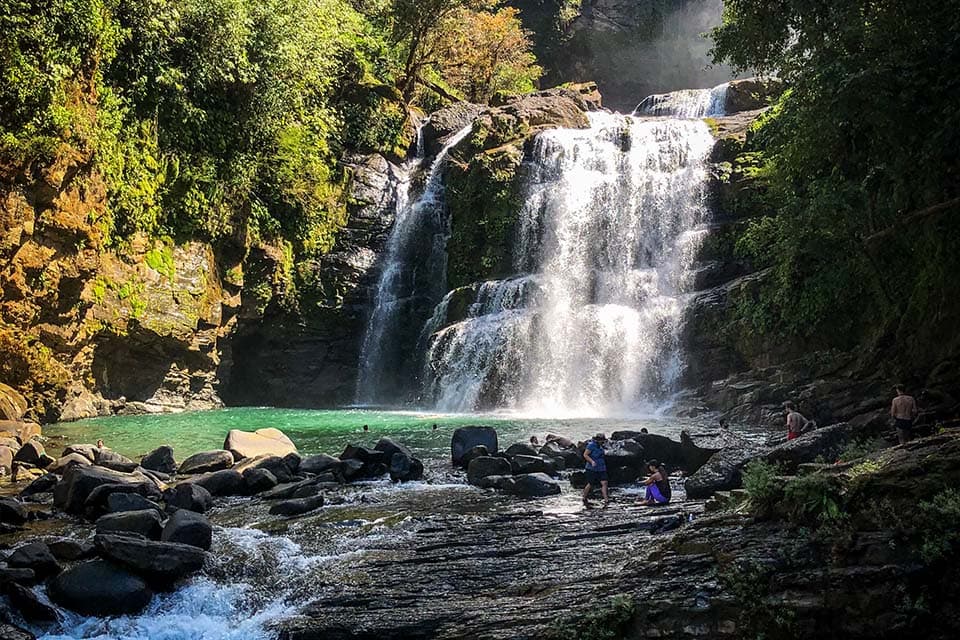 The lower falls and its green pool, with people picking their way across the rocks in front.