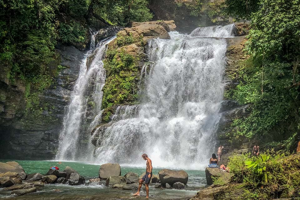 Swimmers on the rocks at the foot of the falls, one walking out across the shallows.