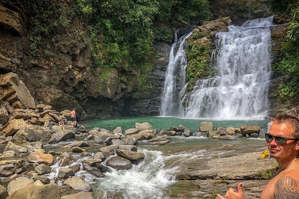 The pool from the far bank, a rope hanging beside the falls and swimmers on the rocks.