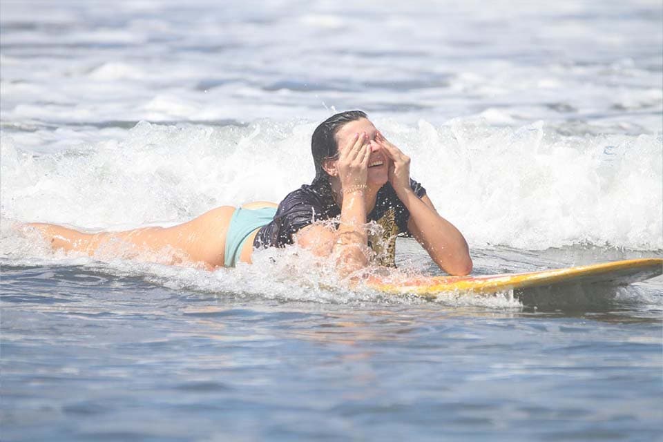 A surfer paddling back out, laughing, with water streaming off her face.