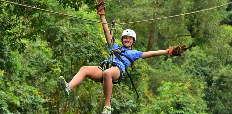 A rider hanging from the pulley mid-line, one arm out, forest filling the frame behind.